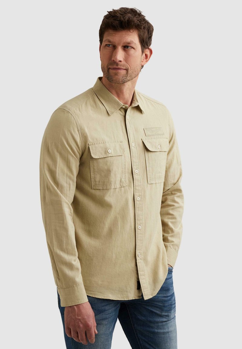 Homme portant une chemise beige à manches longues avec deux poches poitrine et un jean bleu, regardant sur le côté devant un fond gris.