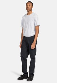 T-shirt en coton gris avec une poche sur le devant, associé à un pantalon en coton noir. Le modèle porte des chaussures noires. Design simple et décontracté, sans motifs notables.