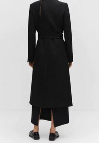 Cappotto lungo nero con vita aderente, realizzato in tessuto testurizzato. Presenta maniche lunghe, una cintura e uno spacco posteriore per facilitare i movimenti.