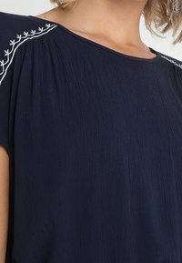 Blouse bleu marine en tissu texturé, avec des manches courtes et des détails brodés blancs le long des coutures des épaules.