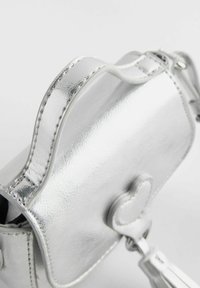Borsa a tracolla metallica argento con design dolcemente curvato, chiusura a forma di cuore e dettaglio con nappina. Texture liscia con cuciture visibili.