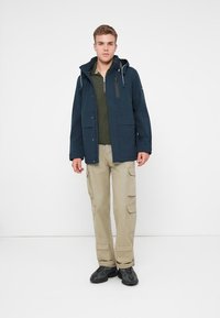 Ναυτικό αδιάβροχο μπουφάν με κουκούλα, εμπρόσθιες τσέπες, φορεμένο πάνω από μια ελαιοπράσινη πλεκτή μπλούζα, σε συνδυασμό με χακί cargo παντελόνια και μαύρα παπούτσια.