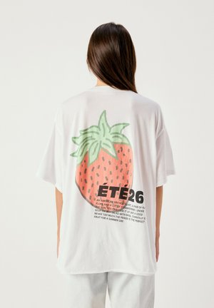 Persona con lunghi capelli castani che indossa una t-shirt bianca oversize con una grande grafica di fragola e testo sul retro.