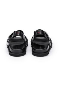Schwarze Ledersandalen mit verstellbaren Riemen, glatter Textur, offenem Rückendesign und flachen Gummisohlen. Mit einem dezenten roten Akzent.