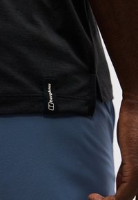 Κοντινό πλάνο ενός ατόμου που φοράει μαύρο πουκάμισο με ετικέτα Berghaus στη ραφή και μπλε παντελόνια, με το χέρι του να ακουμπά δίπλα του.