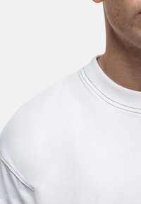 Biała bluza z okrągłym dekoltem, z delikatnymi szwami i miękką teksturą. Materiał wydaje się lekki i lekko rozciągliwy.