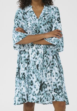 Vrouw draagt een losse, knielange jurk met turquoise en zwarte abstracte luipaardprint en driekwart wijd uitlopende mouwen, armen gekruist.
