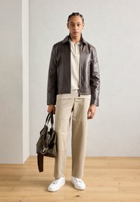 Brun læderjakke, beige tekstureret sweater, cremebukser med syninger, hvide sneakers, holder en grøn taske med mørke detaljer.