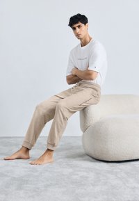 Pantaloni de trening bej cu manșete la gleznă, având un logo pe lateral, asortați cu un tricou alb. Covor texturat de un gri deschis și mobilă moale.