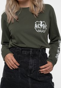 Long-sleeve dark green shirt featuring white AC/DC logo on chest and sleeves, paired with high-waisted black denim skirt.
