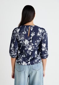 Marinebluse mit weißem Blumenmuster, kurzen Puffärmeln und einem Rückendetail in Form eines Schlitzes. Hergestellt aus leichtem Stoff, kombiniert mit hellen Jeansshorts.