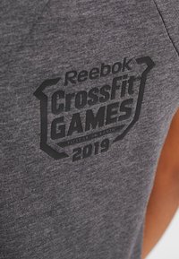 Šedé tričko z látky s černým potiskem loga s nápisem "Reebok CrossFit Games 2019" doplněné geometrickými akcenty a detailním texturováním.