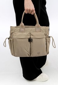 Person hält eine beige Tragetasche mit zwei vorderen Reißverschlusstaschen, seitlichen Kordelzügen und Stoffgriffen, trägt schwarze Hosen und weiße Turnschuhe.