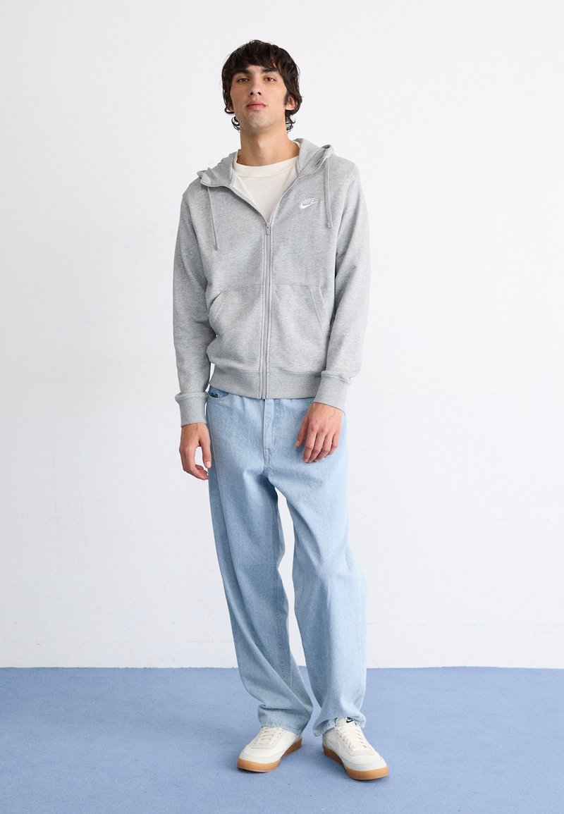 Sudadera gris con cremallera y bolsillo canguro, combinada con jeans de corte holgado en azul claro y zapatillas blancas con detalles en tono marrón. Branding mínimo visible.