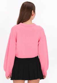 Sweter croppowany w jasnoróżowym kolorze, o dzianinowej fakturze, z długimi rękawami i prążkowanymi mankietami, zestawiony z czarną plisowaną spódnicą.