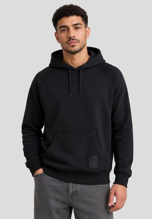 Junger Mann mit lockigem Haar, der einen schwarzen Hoodie und graue Jeans trägt, steht mit einer Hand in der Tasche vor einem schlichten Hintergrund.