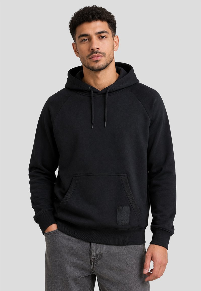 Jeune homme aux cheveux bouclés portant un sweat à capuche noir et un jean gris, debout avec une main dans la poche devant un fond uni.