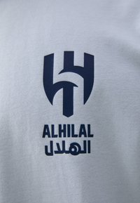 Balta auduma ar tumši zilu logo, kas veidots no stilizētiem burtiem, veidojot vairoga formu, ar arābu tekstu zem tā.