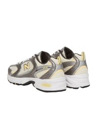 Zapatillas de running de malla en gris y amarillo con detalles en plateado, con un collar acolchado y suelas intermedias blancas. Presentan un prominente logo "N" y una superficie texturizada.