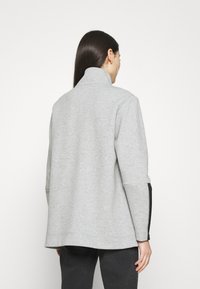 Ljusgrå pulloverjacka med hög krage, sidofickor med dragkedja och en mjuk textur. Har en avslappnad passform och långa ärmar.