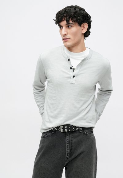 Pilkas ilgomis rankovėmis henlio tipo marškinėlis su sagų juosta, dėvimas ant baltos palaidinės, derinamas su tamsiais džinsais ir juodu diržu su sidabrinėmis akutėmis.