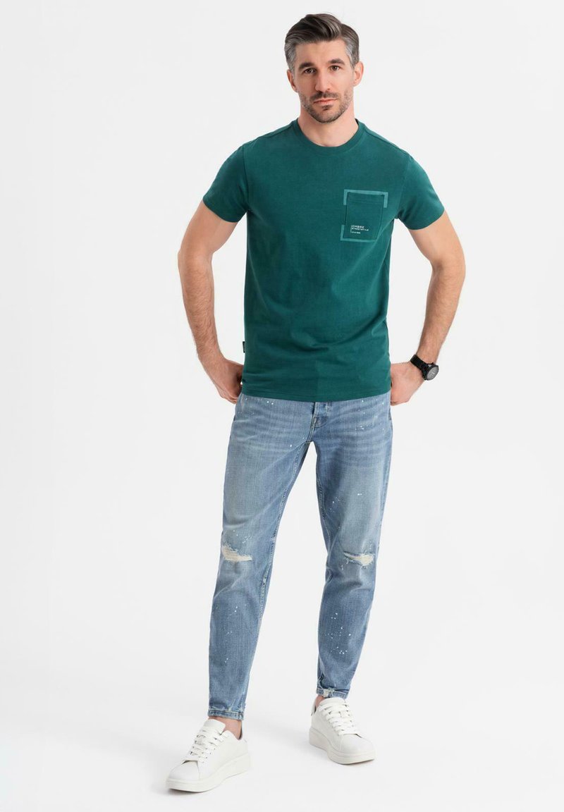 Grön bomullströja med bröstficka och grafisk detalj, ihop med ljusblå, slitna jeans och vita sneakers.