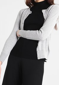 Gråstickad cardigan med mjuk textur, rund halsringning, långa ärmar och pärlknappdetaljer, lagd över en svart ribbad topp.