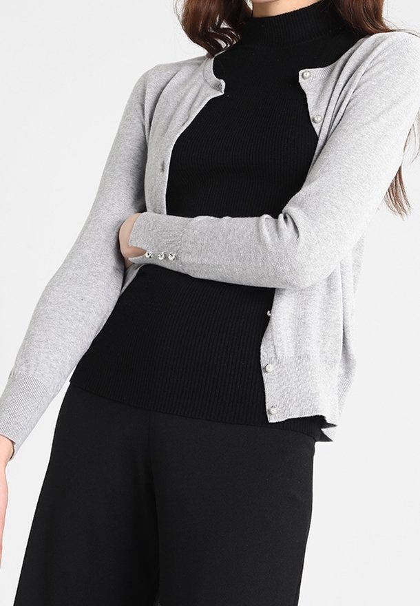 Gråstickad cardigan med mjuk textur, rund halsringning, långa ärmar och pärlknappdetaljer, lagd över en svart ribbad topp.