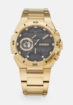 Montre dorée avec un bracelet en métal brossé, cadran noir présentant des indicateurs de jour et de date, des sous-cadrans et de grands index horaires en contraste.