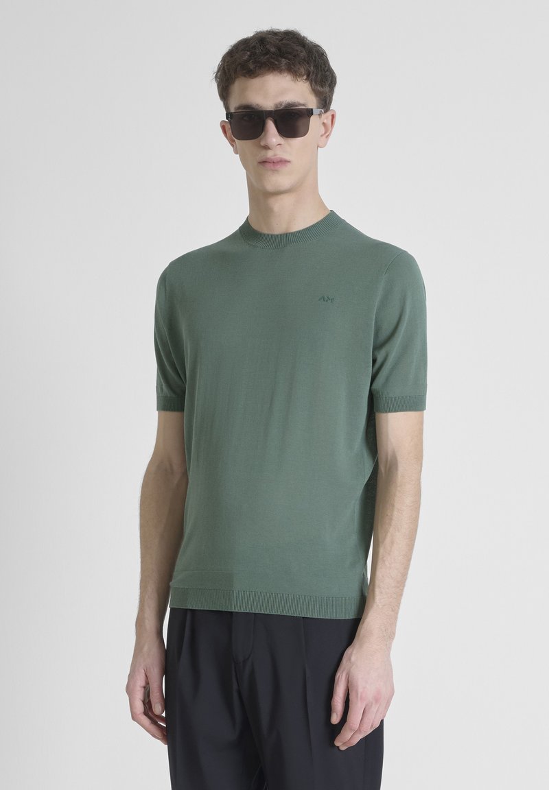 Suéter de punto verde de manga corta con cuello redondo y puños acanalados. Presenta un sutil logo en el pecho. El modelo lleva gafas de sol negras y pantalones.