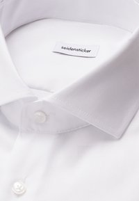 Camicia bianca con colletto classico, realizzata in tessuto morbido. Presenta un orlo cucito e un'etichetta con la scritta "seidensticker".