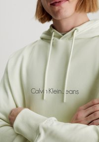 Felpa verde chiaro realizzata in tessuto morbido, con logo frontale "Calvin Klein Jeans" in grigio scuro, polsini a costine e cappuccio regolabile con cordino.