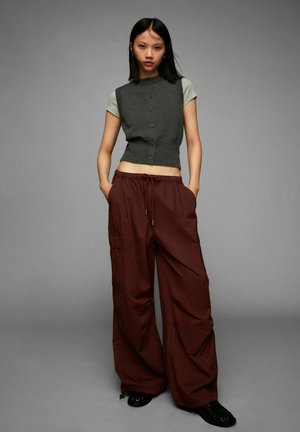 Jeune femme se tient avec les mains dans les poches, portant un haut gris à manches courtes superposé et un pantalon cargo large bordeaux avec des chaussures noires.