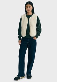 Ung kvinde står iført en cremefarvet vest med knapper over en mørkegrøn sweatshirt, mørkeblå jeans og sorte sneakers, neutral baggrund.