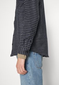Camicia di flanella a quadri blu navy e grigia con maniche lunghe, bottoni ai polsini e un leggero orlo a coda; indossata sopra un maglione beige.