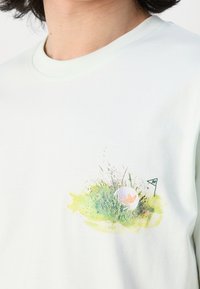 Ljusgrön bomulls t-shirt med en grafisk bild av gräs, en golfboll och en flagga på framsidan. Rund halsringning och korta ärmar.