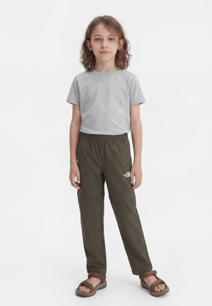 Barn med skulderlangt hår står iført en lys grå kortermet skjorte, olivengrønne bukser med elastisk midje og brune sandaler.
