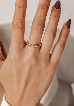 Gouden ring met een gladde, afgeronde vorm, gedragen aan een hand met manicured nagels. De ring heeft een subtiele draaiing in het ontwerp.