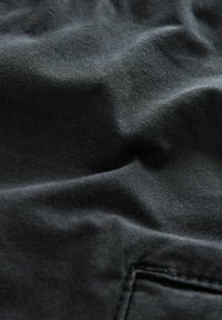 Schwarzer Stoff mit einer weichen, strukturierten Oberfläche, die sanfte Falten zeigt. Verfügt über ein kleines Taschen-Detail in der linken unteren Ecke.