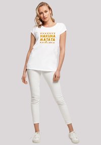 Valkoinen lyhythihainen T-paita, jossa on rohkealla keltaisella tekstillä "HAKUNA MATATA", ympärillä kuvioitu reuna. Yhdistetty valkoisiin slim-fit-housuihin ja tennareihin.
