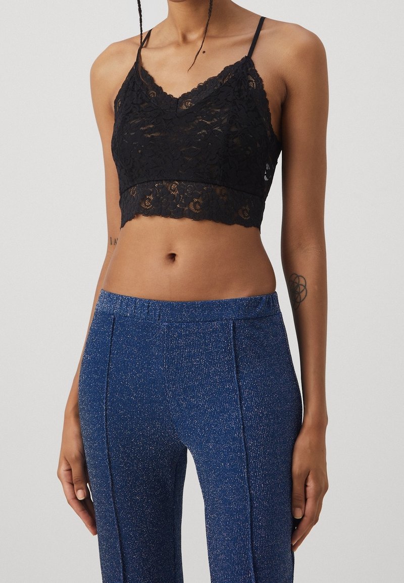 Femme portant un caraco court en dentelle noire et un pantalon bleu texturé taille haute, montrée des épaules aux hanches sur un fond uni.
