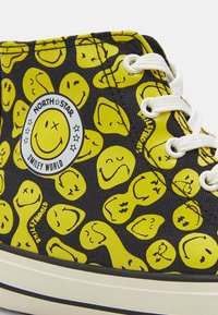 Crne platnene tenisice s ponovljenim uzorkom žutih smajlića. Bijele vezice i kružni logo s natpisom "North Star Smiley World."