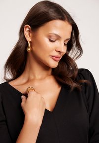 Złote kolczyki w kształcie obręczy oraz pasujący pierścionek są noszone z czarną bluzką z dekoltem w kształcie litery V. Kolczyki mają gładki, zaokrąglony kształt i błyszczące wykończenie.