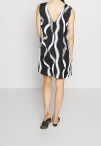 Ärmlös svart klänning med vita vågmönster, V-formad rygg, knälång, slät tyg, kombinerad med svarta sandaler.