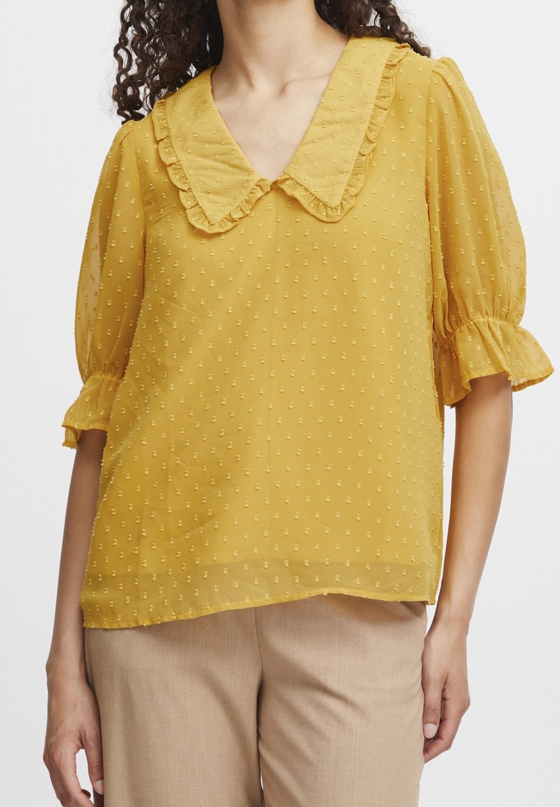 Senfgelbe Bluse mit einem rüschenden Kragen, kurzen Puffärmeln und strukturierten Punkten. Leichtes, halbtransparentes Material.