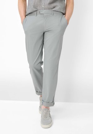 Mand iført lysegrå opkørte bukser, grå sneakers og en lysegrå kortærmet skjorte med hænderne i lommerne.