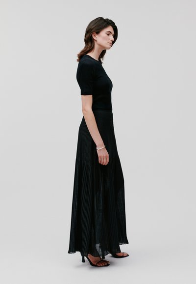 Schwarzer, plissierter Maxirock mit einem enganliegenden schwarzen Oberteil. Der Rock hat vertikale Falten, und das Model trägt Sandalen mit offenem Zehenbereich. Minimalistisches Design.