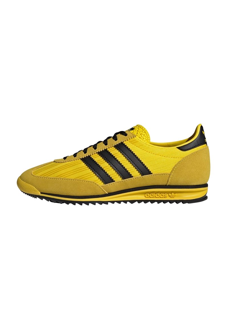 Zapato deportivo amarillo con parte superior de ante y tela, diseño de tres franjas negras, acentos texturizados y suela de goma.
