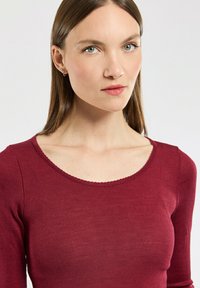 Junge Frau mit glatten braunen Haaren, die kleine Creolen trägt und ein enges, gerippte burgunderfarbenes Langarmoberteil anhat, vor neutralem Hintergrund.