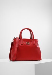 Sac à main en cuir rouge avec des poignées rondes doubles, une bandoulière amovible et un accent logo métallique à l'avant. Texture lisse.
